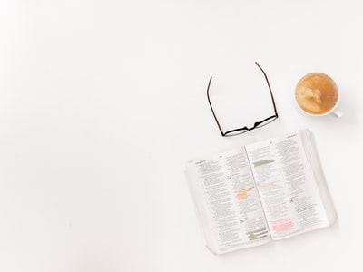 Beside cappuccino cup with a black frame glasses, white surface has printed books
